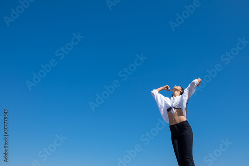 Happy woman with tanned slim body breathing fresh air raising her arms up, enjoying a sunny summer holiday on beach destination against blue sky, outdoors. Travel and well being lifestyle.