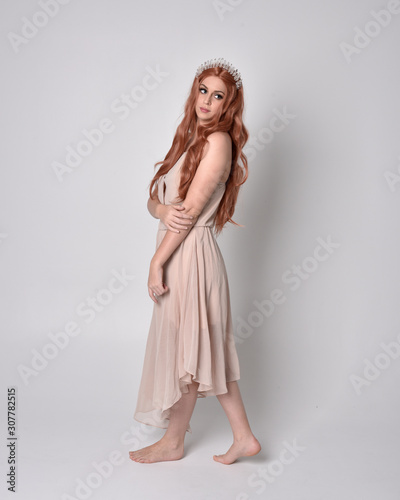 full length portrait of a pretty, fairy girl wearing a nude flowy dress and crystal crown. Standing and dancing pose against a grey studio background.