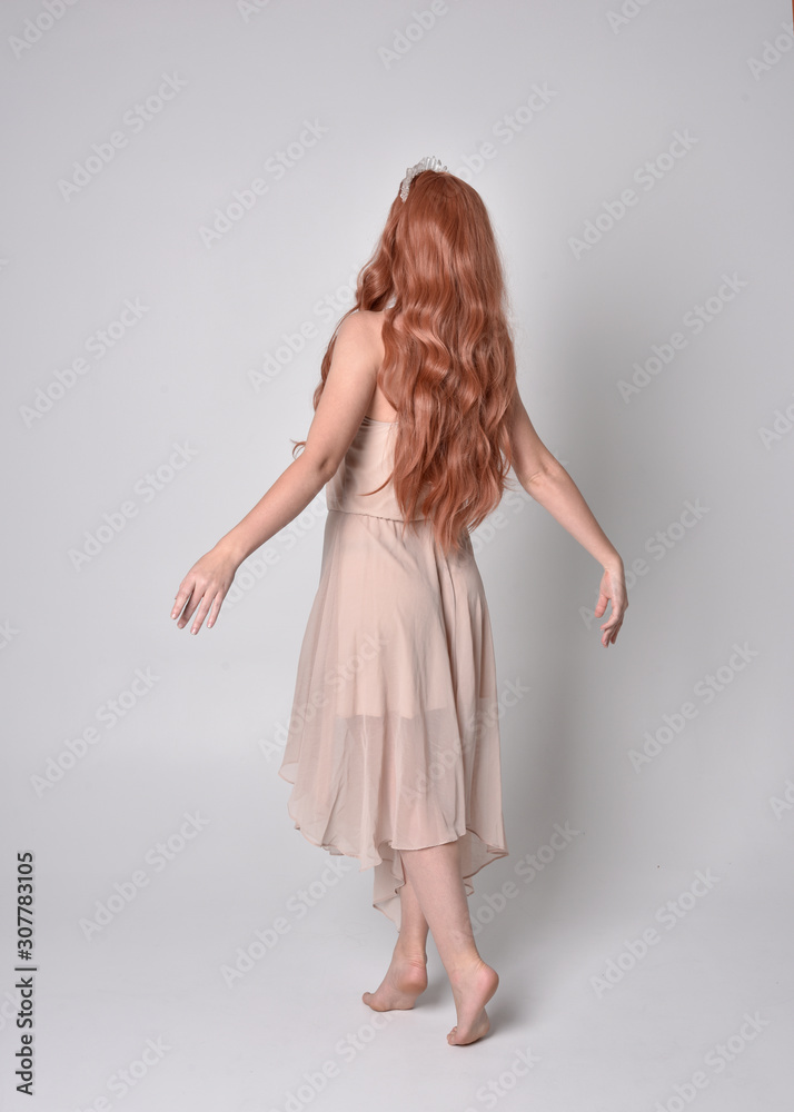 full length portrait of a pretty, fairy girl wearing a nude flowy dress and crystal crown. Standing and dancing pose against a grey studio background.