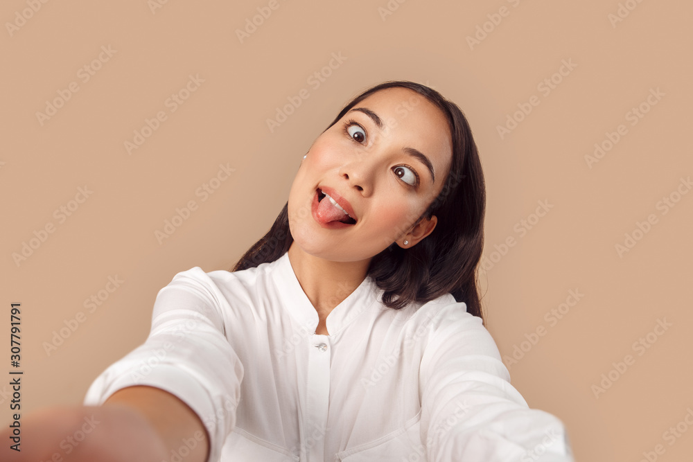 Freestyle. Young woman in shirt standing isolated on bage taking selfie taking selfie on smartphone showing tongue grimacing silly looking at nose close-up Freestyle. Young woman in shirt standing isolated on bage taking selfie taking selfie on smartphone showing tongue grimacing silly looking at nose close-up