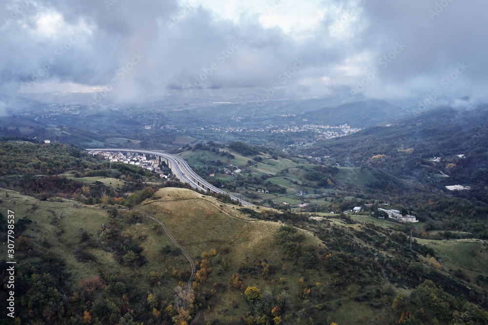 Autumn alpine speed road in Italia. Aerial view from above with clouds on the background and village far away