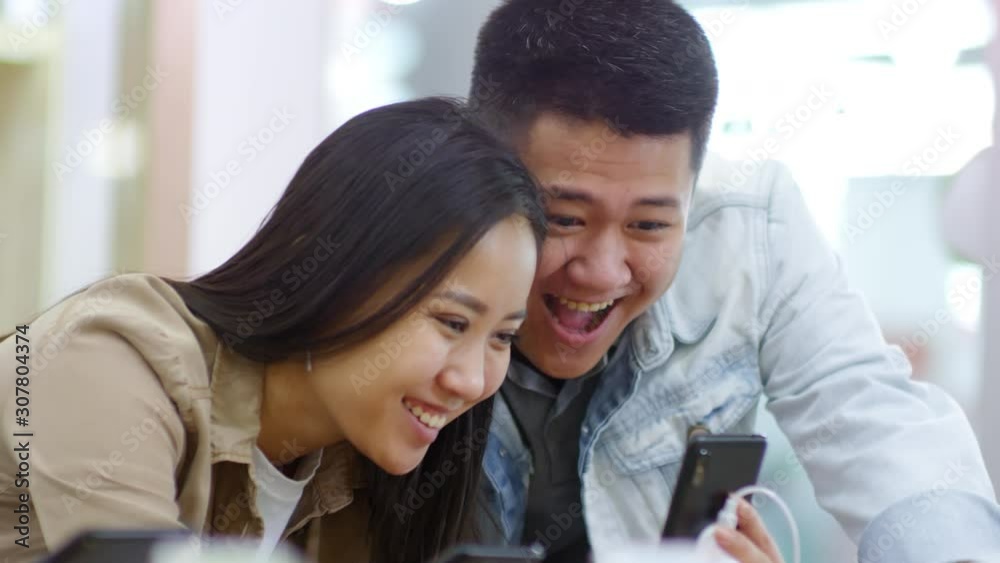 Medium shot of young Asian couple using smartphone touchscreen while choosing phone model at gadget store Medium shot of young Asian couple using smartphone touchscreen while choosing phone model at gadget store