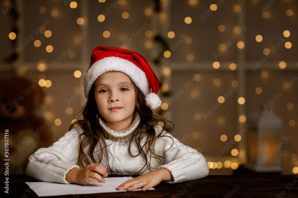 Happy little child girl in santa hat writing wish list or letter to Santa Claus on background with lights. Merry Christmas