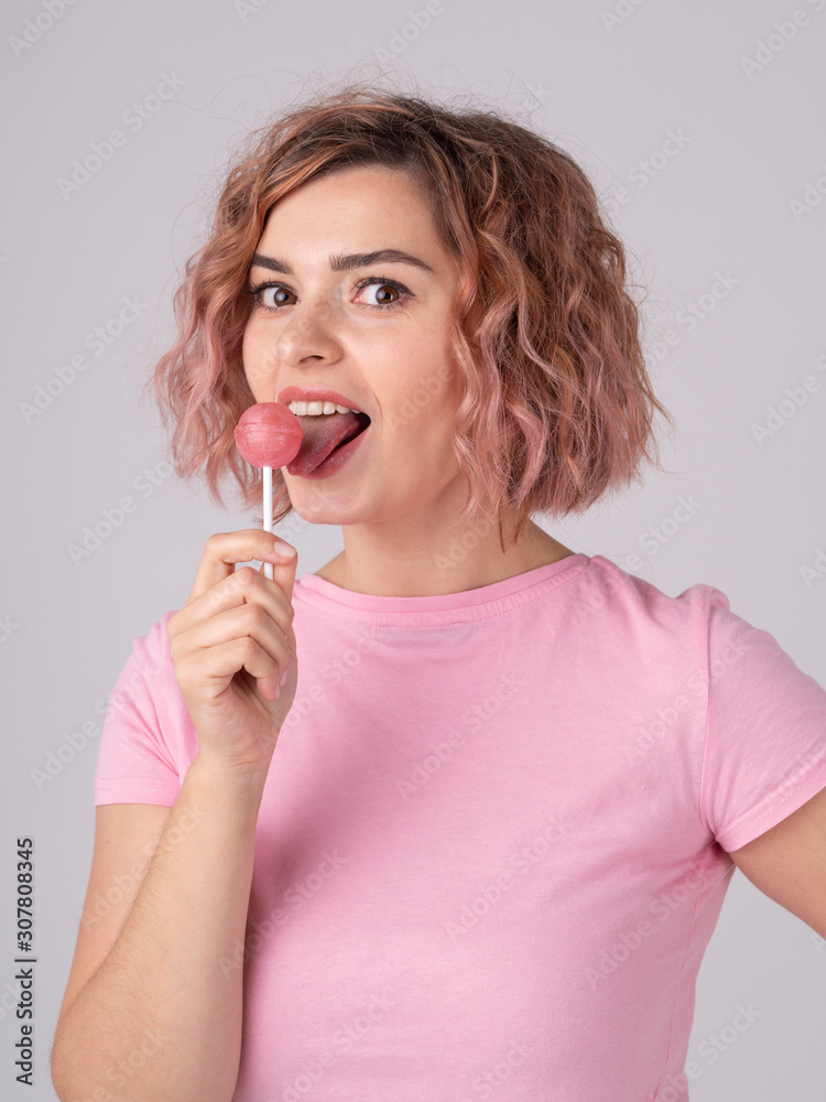 Hot young woman dressed in a pink t-shirt licking pink lollipop looking forward standing on grey background Hot young woman dressed in a pink t-shirt licking pink lollipop looking forward standing on grey background