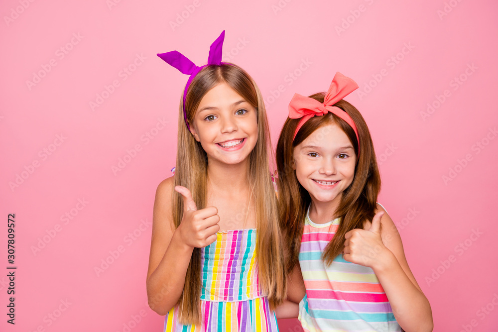 Close up photo of charming kids hugging embracing showing thumb up wearing bright headbands skirt dress isolated over pink background Close up photo of charming kids hugging embracing showing thumb up wearing bright headbands skirt dress isolated over pink background