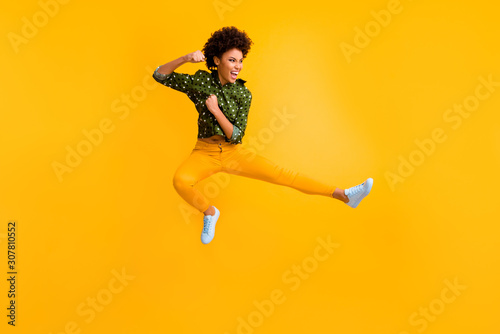 Full length photo of crazy dark skin curly lady jumping high making fighting moves ready to kick leg and fist wear green dotted shirt trousers isolated yellow color background photo