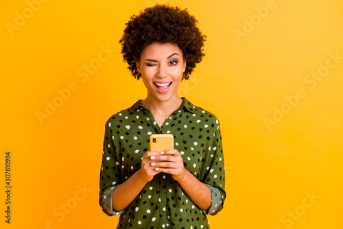 Portrait of excited crazy afro american girl hold smart phone use read social network news wear stylish green bright clothing isolated over yellow color background