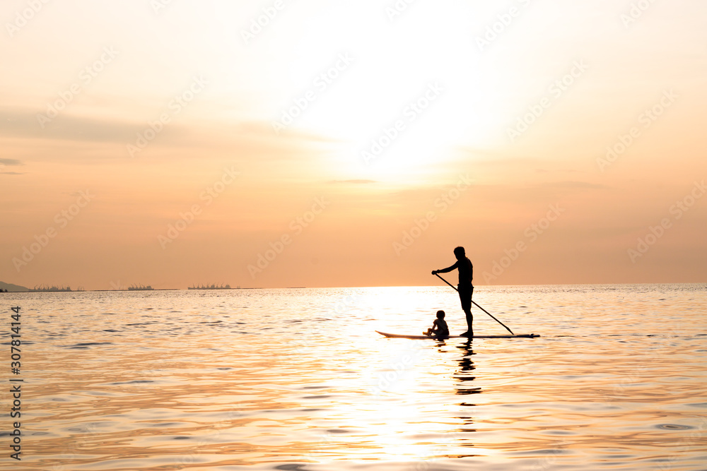 Silhouette of dad and son playing the stand-up paddle board on the sea with beautiful summer sunset colors. Happy family concept.