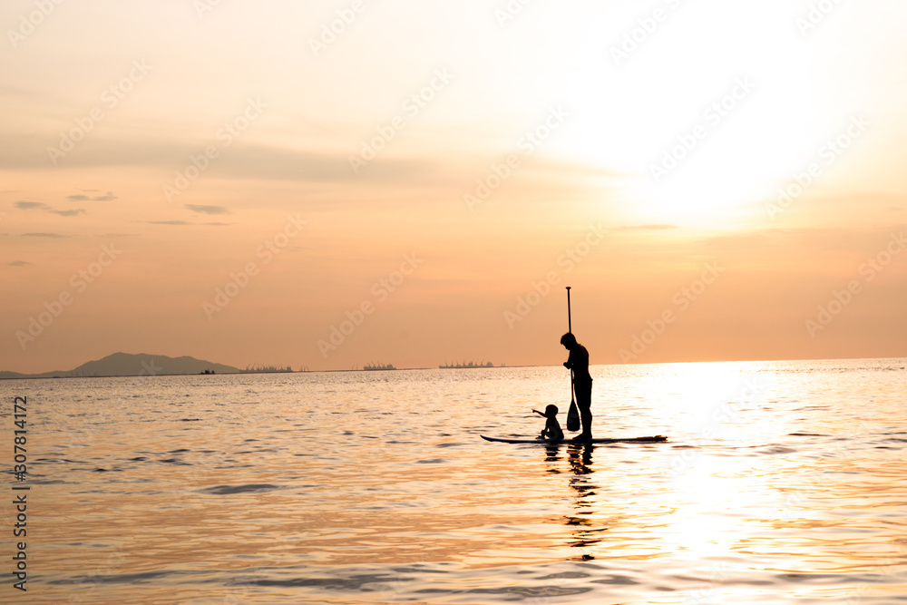 Silhouette of dad and son playing the stand-up paddle board on the sea with beautiful summer sunset colors. Happy family concept.