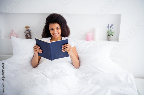 Photo of charming curly dark skin lady lying sheets bed covered white blanket hold book reading novel spend free time useful weekend saturday morning wear pajama room indoors