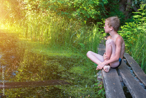 A boy in swimming trunks sits on the edge of a wooden bridge near the water. photo