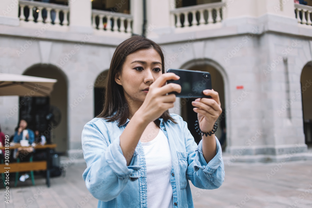 Focused beautiful Asian tourist taking photo with smartphone