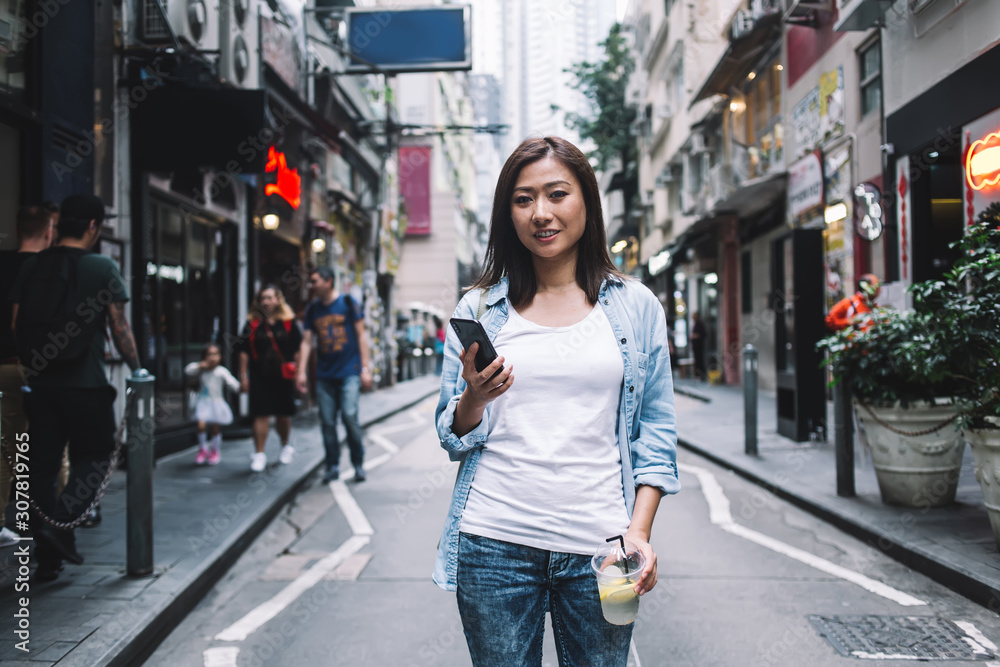 Cheerful Asian woman standing on street with smartphone and drink