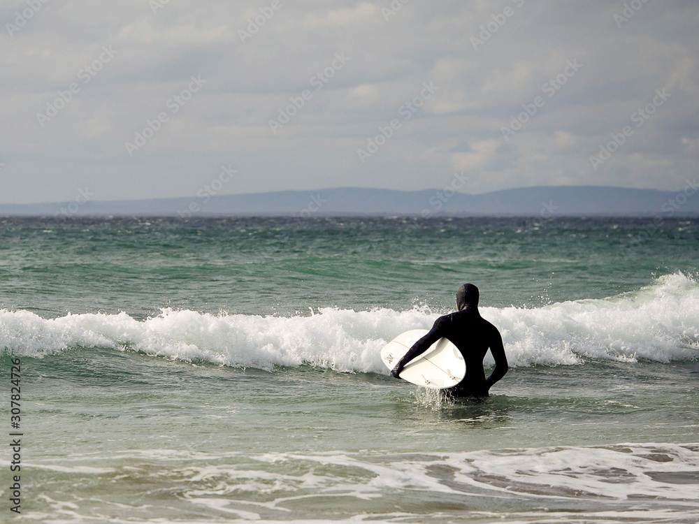 Surfer in black wet suit caring his board under left hand going deep into ocean water. Concept sport, surfing, outdoor activity.