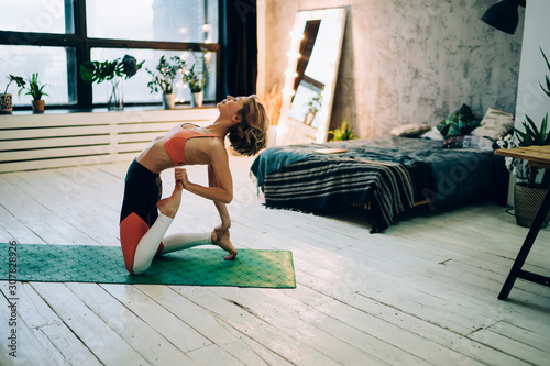 Calm hipster girl stretching back and legs for training body muscles during home workout on green mat, attractive active female athlete doing exercise practicing for stretching flexibility in flat