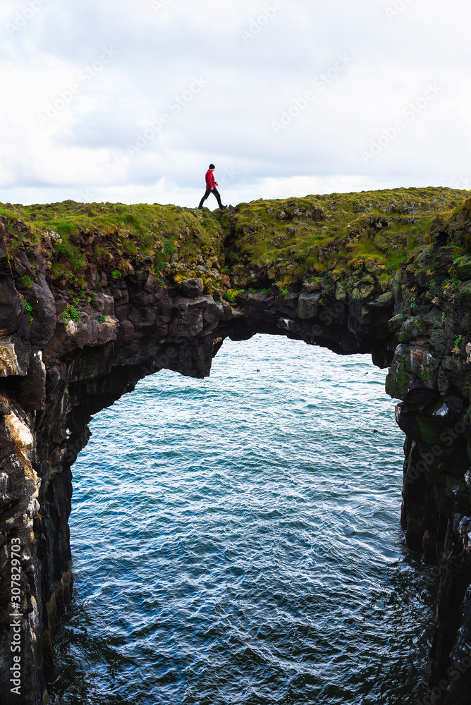 Tourist walks over a natural rock bridge in Arnarstapi, Iceland