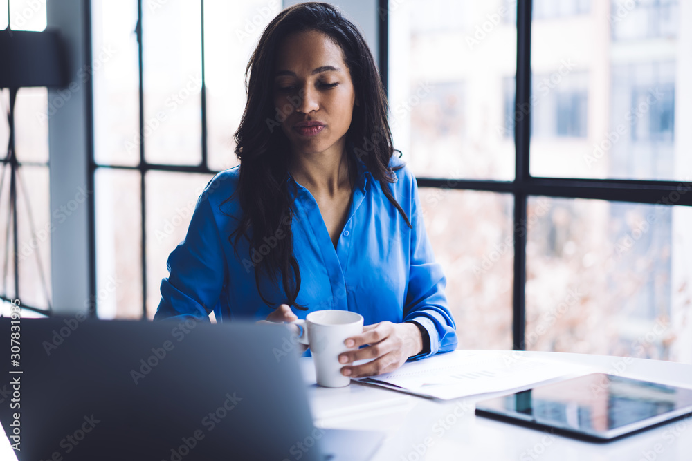 Frown elegant ethnic business lady focusing on laptop screen while sitting and holding cup Frown elegant ethnic business lady focusing on laptop screen while sitting and holding cup