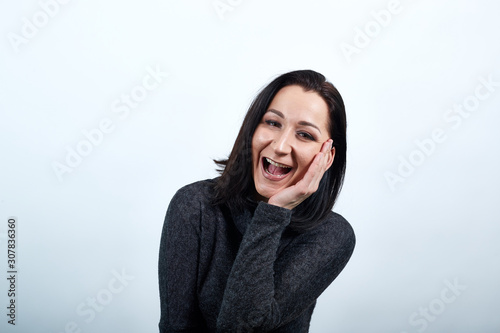 Attrictive smiling young woman in nice black sweater keeping hand on cheek, laughting over isolated white background, Lifestyle concept photo