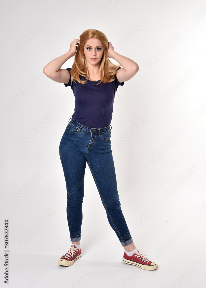 Full length portrait of a pretty blonde girl wearing casual blue shirt, denim jeans and sneakers. Standing pose on a studio background. Full length portrait of a pretty blonde girl wearing casual blue shirt, denim jeans and sneakers. Standing pose on a studio background.