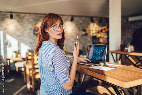 Serious caucasian woman in eyewear look at camera dialling number on mobile phone working remotely in coworking space, businesswoman using technologies for online research and job download updates
