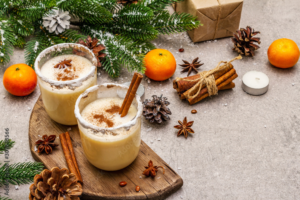 Eggnog with cinnamon and nutmeg for Christmas and winter holidays. Homemade beverage in glasses with spicy rim. Tangerines, candles, gift. Stone concrete background