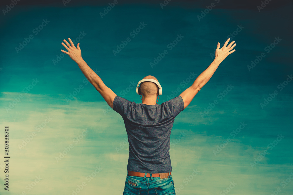 A bald man in headphones listens to music, standing with his back to the camera, raising his hands up on a blue background.