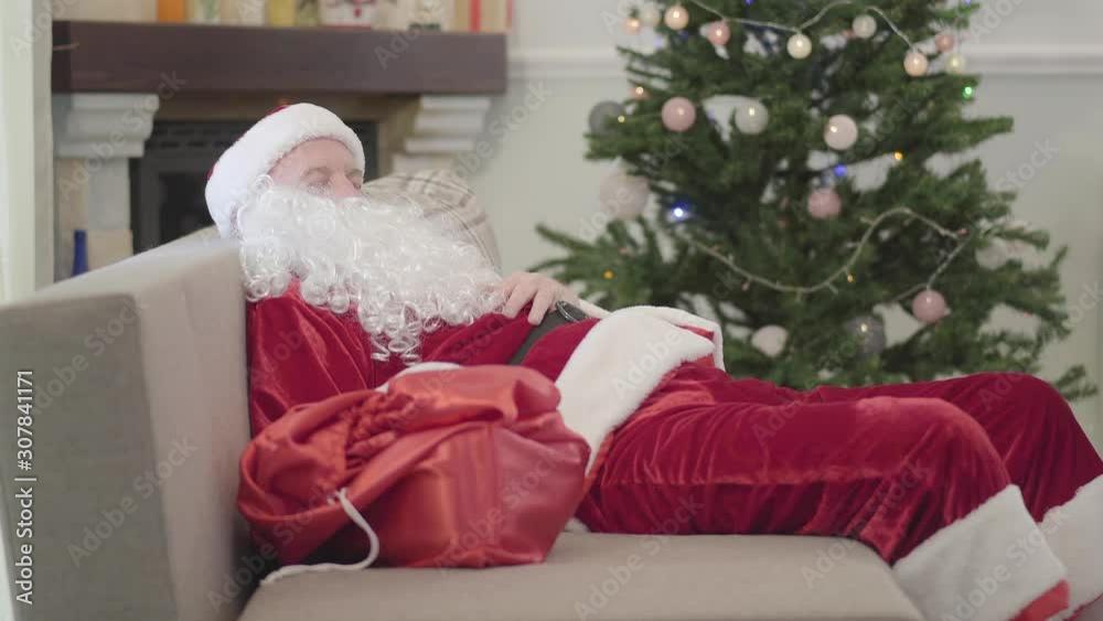 Old Caucasian man in Santa Claus costume sleeping on the sofa at the background of Xmas tree. Exhausted guy resting after difficult working day at home. Old Caucasian man in Santa Claus costume sleeping on the sofa at the background of Xmas tree. Exhausted guy resting after difficult working day at home.