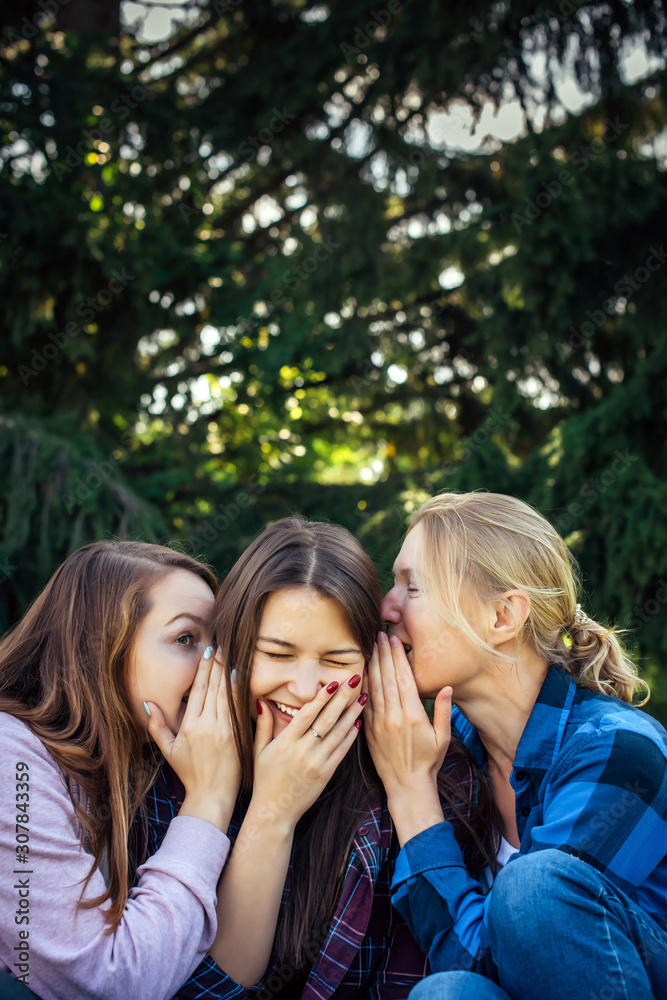 Three cheerful girls whisper and gossip against green foliage in the park. Women joke and laugh, vertical image. Three cheerful girls whisper and gossip against green foliage in the park. Women joke and laugh, vertical image.