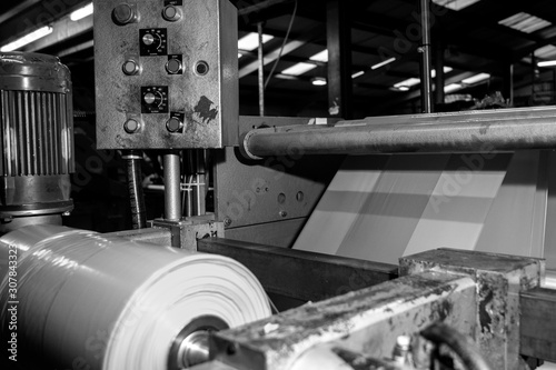 Black & White imagery of spool of plastic packaging sheet heavy duty for industrial use outdoors. wrapped around a metal pole ready to feed into a machine for cutting into shape or printing logos.