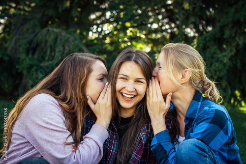 Three young attractive woman sharing secrets sitting on green grass in the park. Cheerful girlfriends gossip and whisper outdoor.