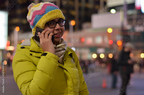 Lifestyle portrait shot of smiling young Indian woman, wearing jacket and cap, speaking on her mobile phone in the American winters. Bokeh of Christmas lighting can be seen as boken in the background