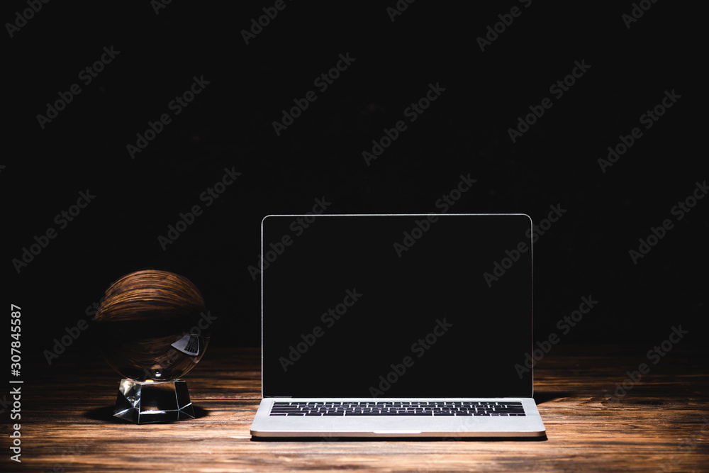 Crystal ball near laptop on wooden table on black background - obrazy, fototapety, plakaty 