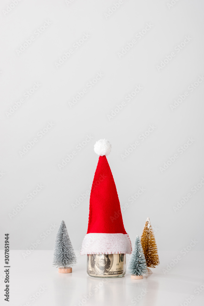 decorative and small pines near jar with dollar banknotes with santa hat on white decorative and small pines near jar with dollar banknotes with santa hat on white