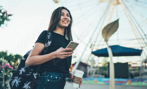 Happy hipster girl traveler using app on smartphone while having trip visiting city festival, cheerful female international student enjoying free time on vacations in amusement park holding cellular. © BullRun