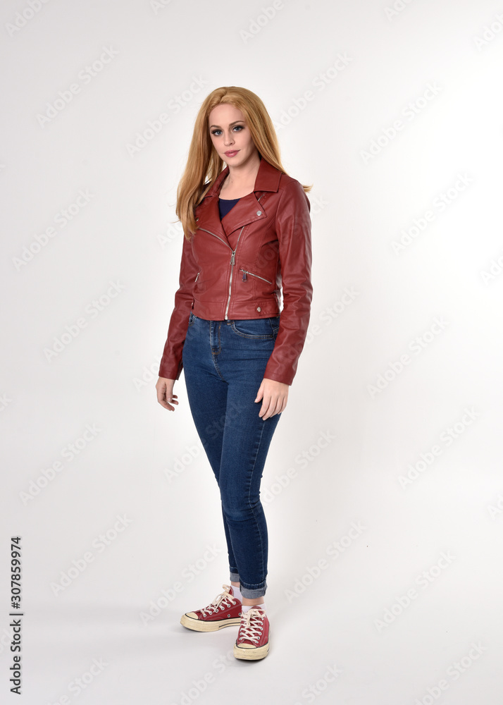 Full length portrait of a pretty blonde girl wearing red leather jacket denim jeans and sneakers. Standing pose on a studio background. Full length portrait of a pretty blonde girl wearing red leather jacket denim jeans and sneakers. Standing pose on a studio background.
