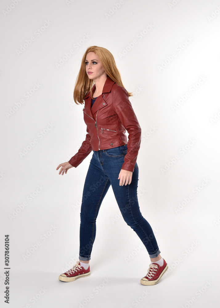 Full length portrait of a pretty blonde girl wearing red leather jacket denim jeans and sneakers. Standing pose on a studio background. Full length portrait of a pretty blonde girl wearing red leather jacket denim jeans and sneakers. Standing pose on a studio background.
