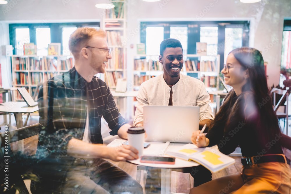 Group of diverse students sitting in coffee shop and laughing during time for cooperation and learning indoors, happy successful millennials discussing ideas and smiling near table in college campus Group of diverse students sitting in coffee shop and laughing during time for cooperation and learning indoors, happy successful millennials discussing ideas and smiling near table in college campus