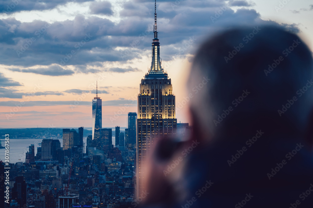 Obraz premium Back view of male traveler taking photo of skyscrapers in skyline at evening time exploring New York city during holidays. Sightseeing to Famous Empire State Building Landmark. Scene cityscape view