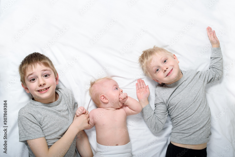 Two older brothers are lying with the youngest baby in a white bed. Happy childhood, big family Two older brothers are lying with the youngest baby in a white bed. Happy childhood, big family