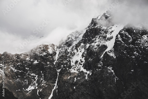 Atmospheric minimalist alpine landscape with snowy rocky mountain peak. Low clouds near snowbound range. Rocks with snow in mist. Craggy mountains in fog. Majestic misty foggy scenery on high altitude © Daniil
