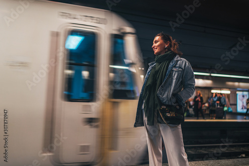 Elegant woman dressed in oversize denim jacket and wide white trousers waiting for a subway train which arriving right now
