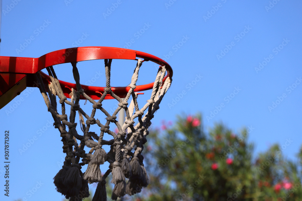 Red basketball basket on a background of blue sky and trees on a sunny day. Cropped shot, horizontal, close-up. Sport and hobby concept.