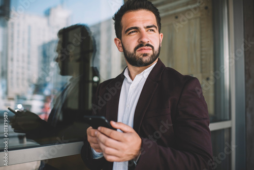 Pensive caucasian man in formal outfit holding smartphone pondering on smth planning to send message, serious male in trendy jacket looking away using mobile phone and 4G for browsing outdoors.