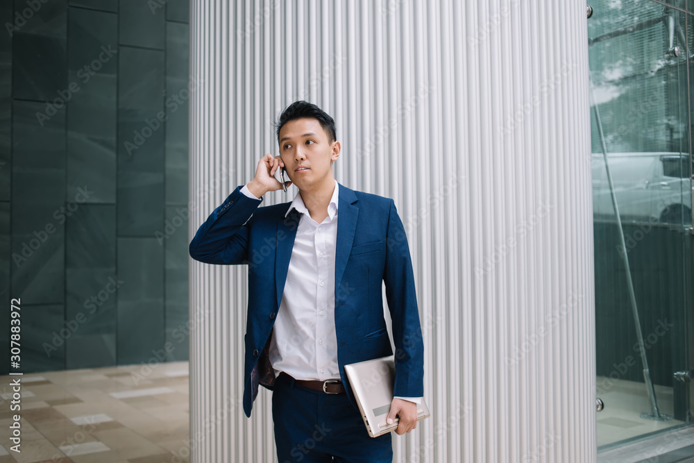 Professional young modern Asian businessman with laptop standing and speaking on smartphone