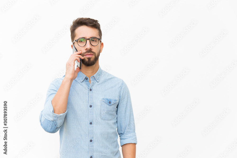 Pensive focused guy speaking on cellphone, having serious phone talk. Handsome young man in casual shirt and glasses standing isolated over white background. Communication concept Pensive focused guy speaking on cellphone, having serious phone talk. Handsome young man in casual shirt and glasses standing isolated over white background. Communication concept