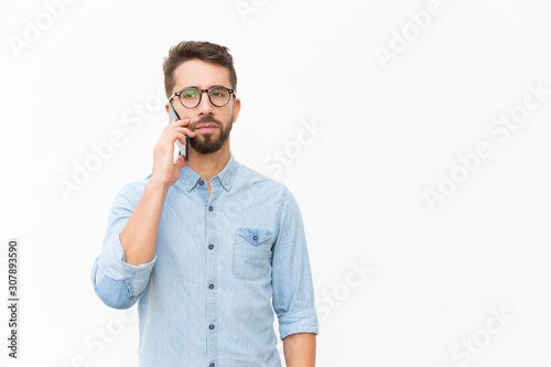 Pensive focused guy speaking on cellphone, having serious phone talk. Handsome young man in casual shirt and glasses standing isolated over white background. Communication concept