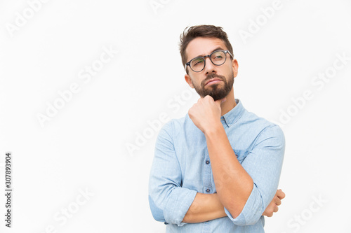 Pensive male customer looking away at copy space, touching chin, thinking. Handsome young man in casual shirt and glasses standing isolated over white background. Advertising concept