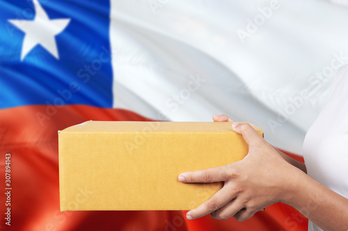 Chile delivery service. International shipment theme. Woman courier hand holding brown box isolated on national flag background. © sezerozger