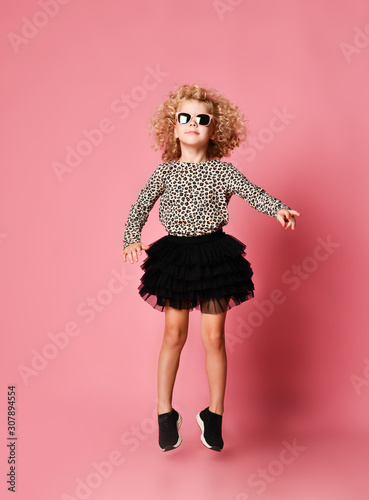 Blonde kid girl with curly hair in leopard print shirt and fatin skirt is jumping flying, dancing on pink photo