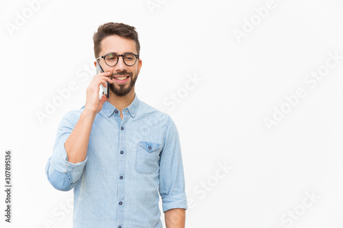 Positive guy talking on cellphone, enjoying nice conversation. Handsome young man in casual shirt and glasses standing isolated over white background. Phone call concept